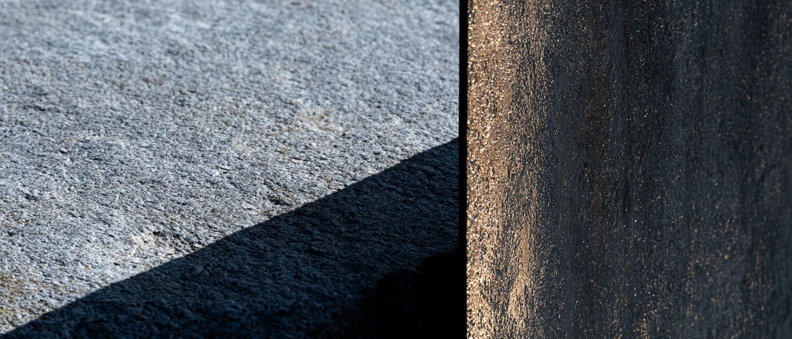 Close detail of weathered grey granite meeting darkened bronze, lit from one side.