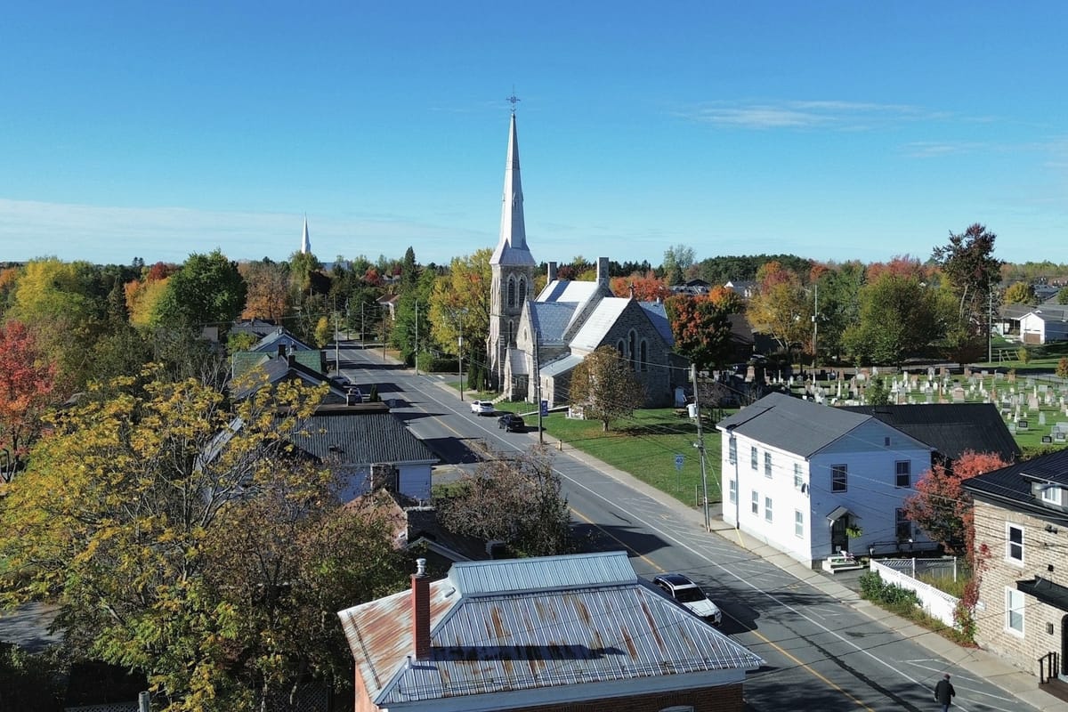 A small eastern Ontario village from above on an autumn afternoon: a stone church and spire on a two-lane main street, clapboard houses, a churchyard and maples turning colour.
